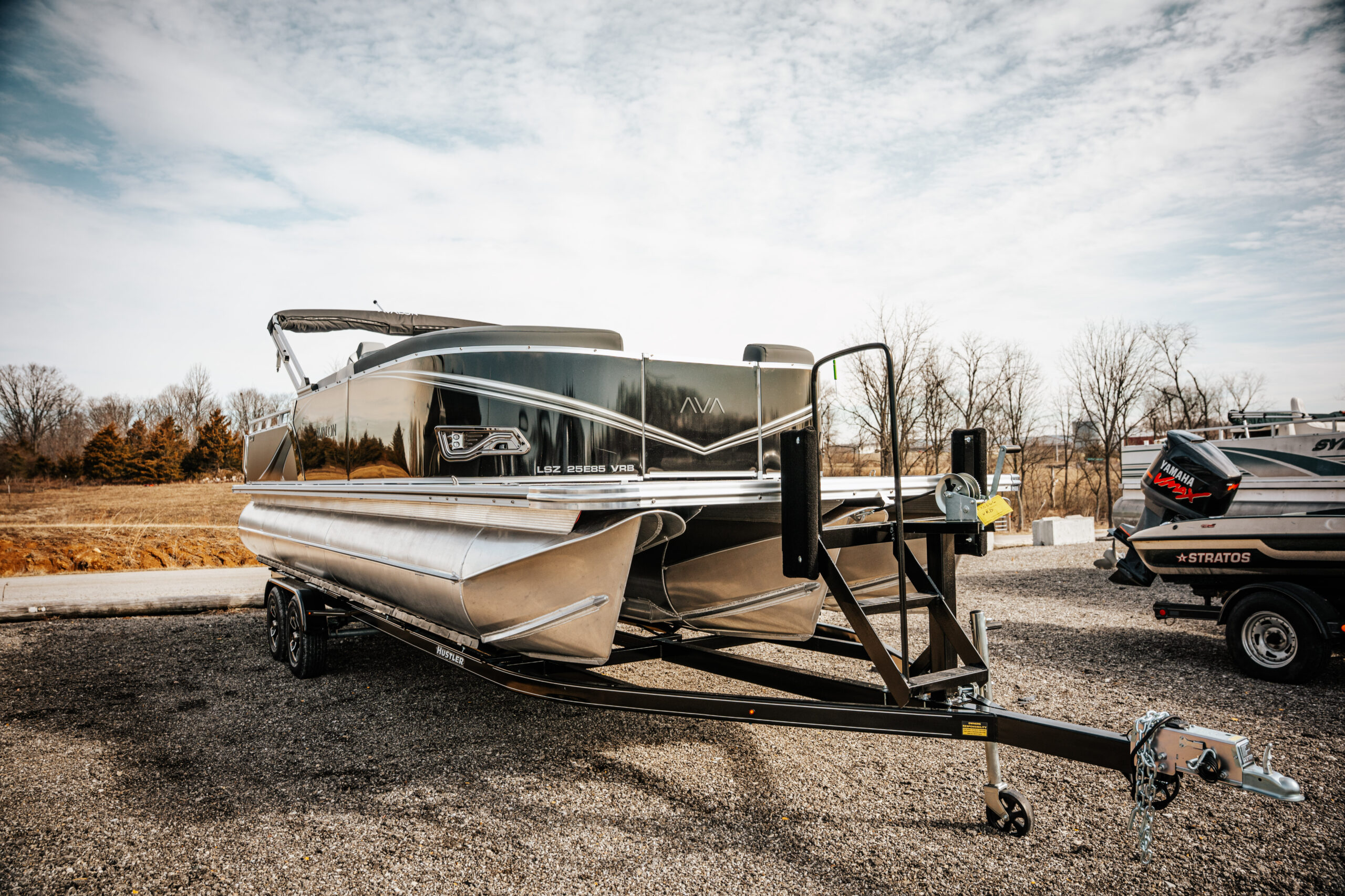 2026 Avalon LSZ boat in flat black and gloss black colors on a trailer, showcasing sleek design and aluminum pontoons, set against a clear sky and gravel parking area.