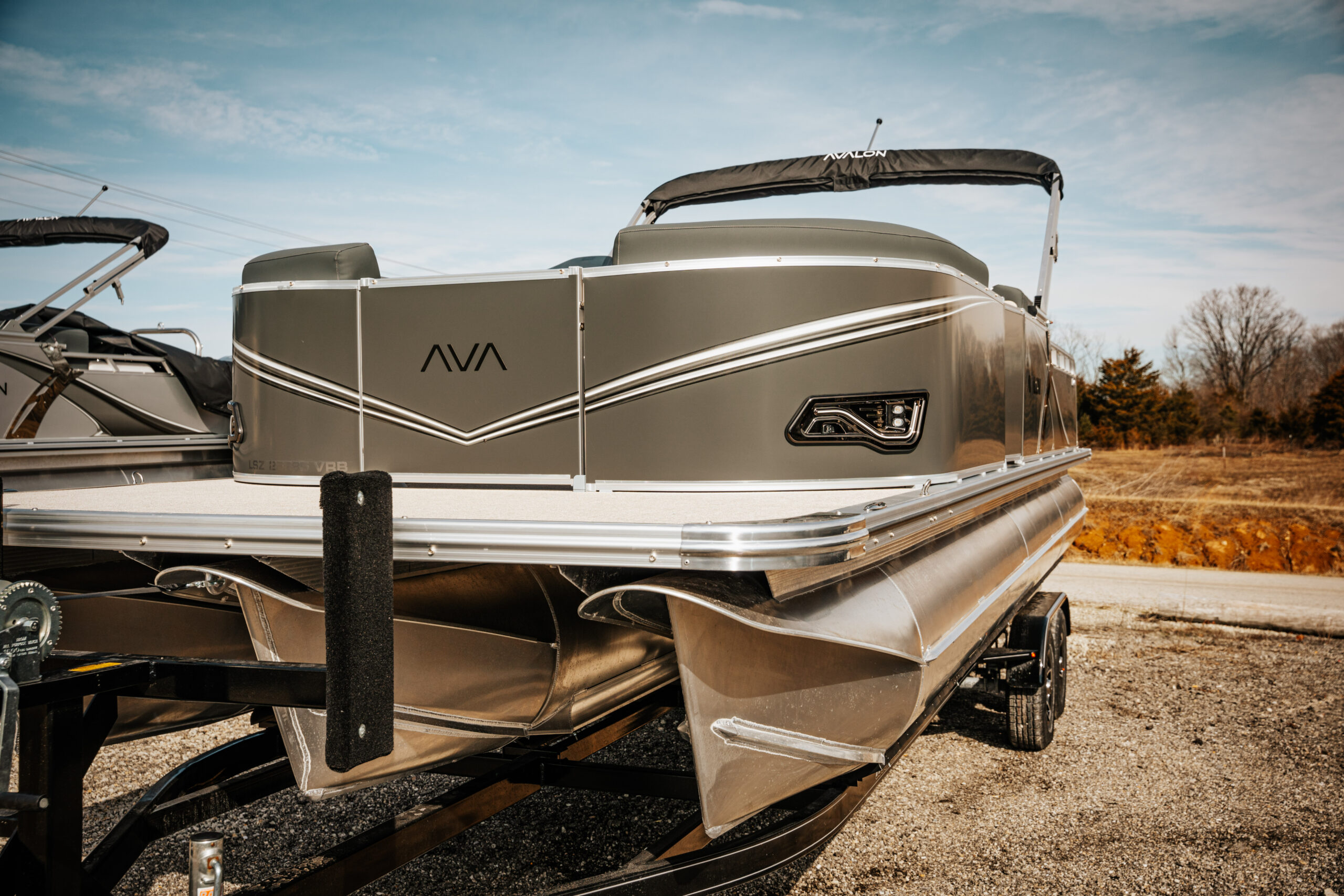 Avalon LSZ pontoon boat in silver metallic, showcasing sleek design and modern features, parked on a trailer against a clear blue sky.