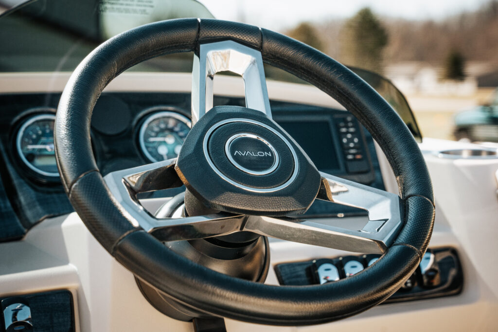 Steering wheel of the 2026 Avalon Catalina Quad Lounger with Avalon logo, featuring control panel and gauges in a matte black interior setting.