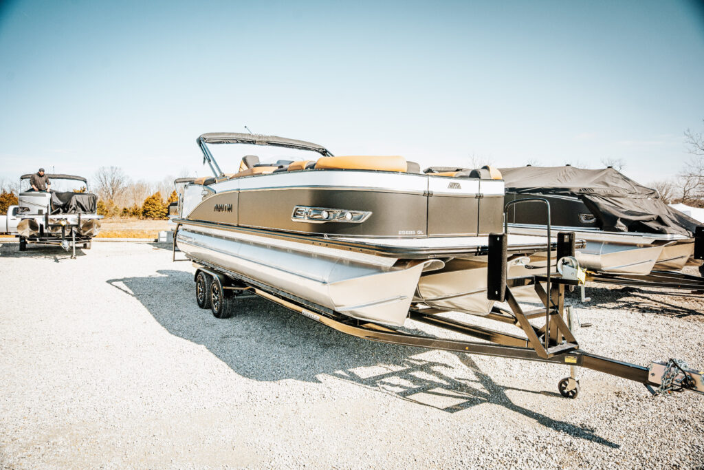Avalon pontoon boat on trailer, featuring a sleek design with black and silver accents, parked in a marine lot, with another boat in the background, showcasing marine sales options.