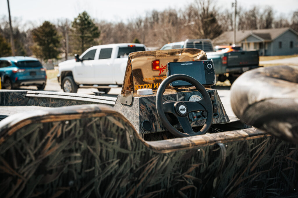 Dashboard of the 2024 Polar Kraft Sport X 190 SC boat featuring a steering wheel, fish finder, and camouflage design, with parked vehicles and trees in the background.