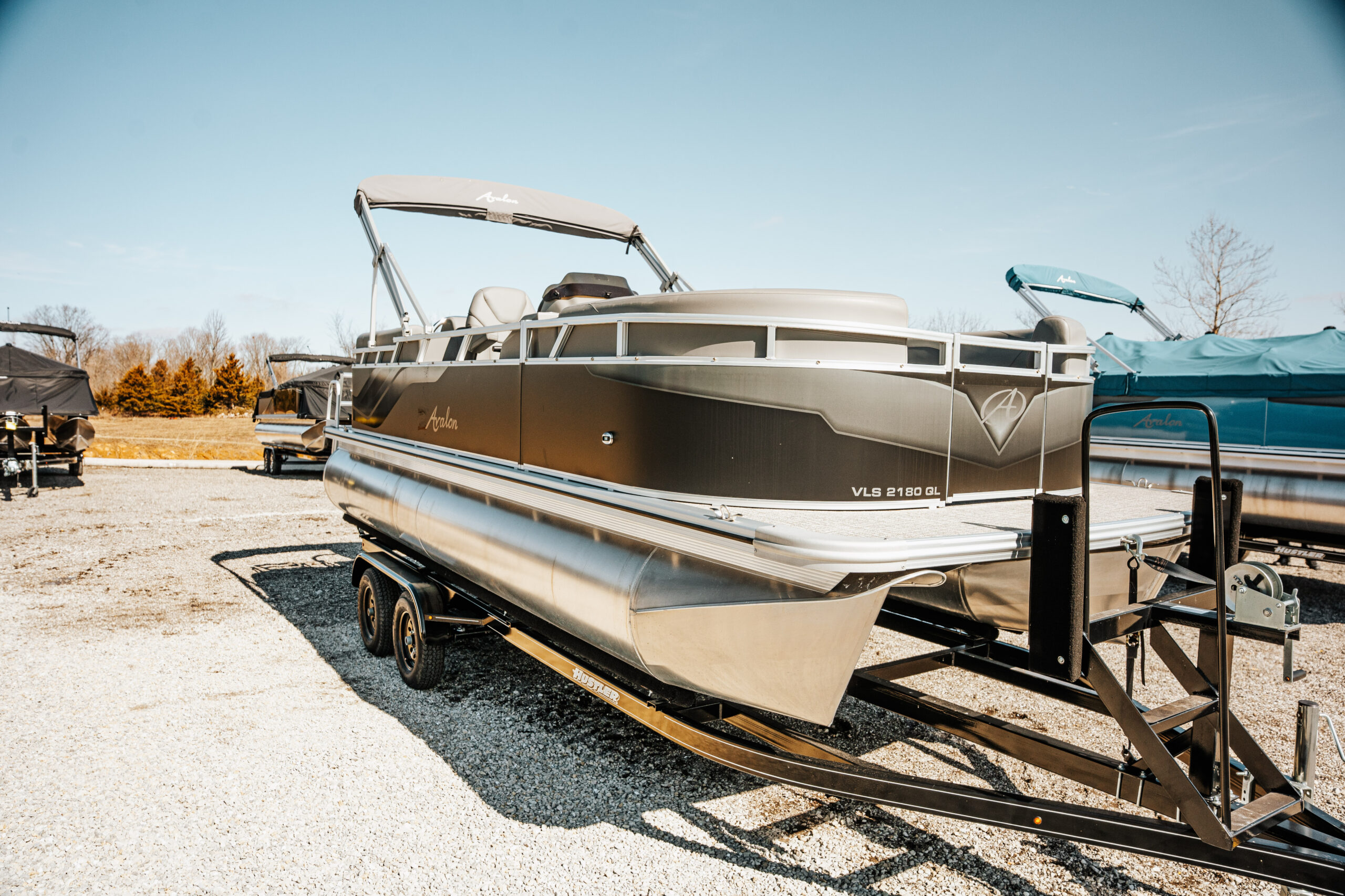 2024 Avalon Venture Quad Lounger boat on trailer, showcasing sleek design and features, with surrounding boats and clear blue sky in the background.